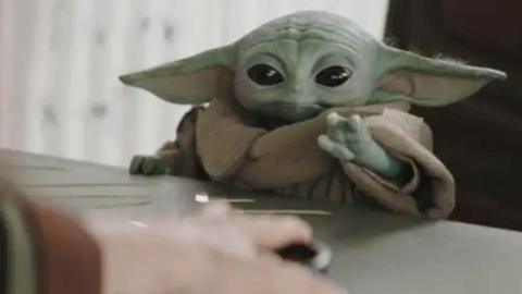 Baby Yoda - a green little space creature - in a sort of gown out stretches his left arm reaching for something on a table. You can see the hand of a person in the foreground who appears to be passing the item over to Baby Yoda