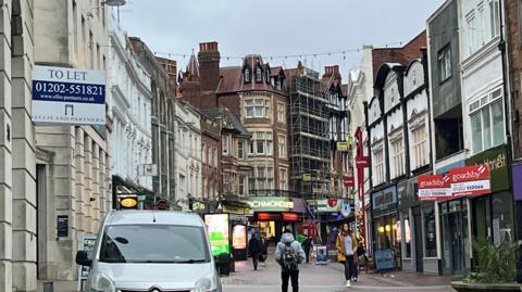 A high street with people walking. To let and signs above some of the empty shops on the high street. 