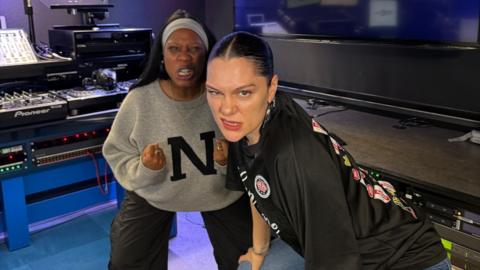 Jessie J and Nadia Jae make faces while crouching in a radio studio