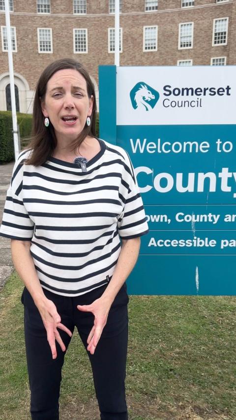 Reporter Ruth Bradley stands in front of a Somerset Council sign welcoming people to County Hall in Taunton. County Hall is visible in the background. Ruth has shoulder-length brown hair. She is wearing black trousers and a short-sleeved top with white and black horizontal stripes. She has circular cream, green and pink coloured earrings with lightning bolts on. 