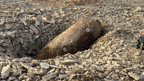 A large, rusted cylindrical object, a a German SC250 air-dropped bomb, lying on its side in a shallow pit at what appears to be a construction or excavation site. The object is partially embedded in loose grey rubble made up of broken stone and aggregate.