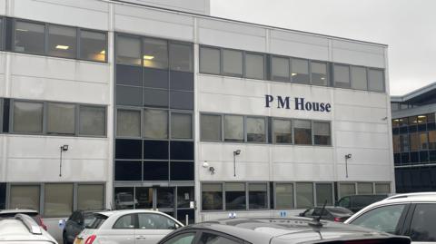 The image shows a modern, three‑storey office building with a mostly white and grey exterior. Large rectangular windows line each floor, with some panels appearing darker than others, giving the façade a slightly varied pattern. The building has the name “PM House” displayed prominently in dark blue lettering on the front.
Several cars are parked in the foreground.