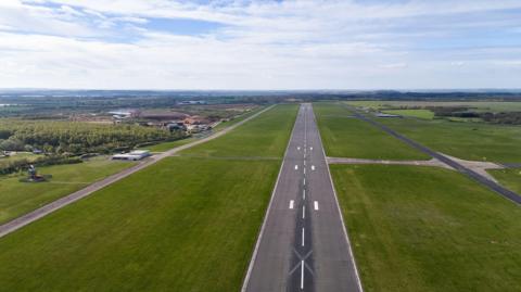 Aerial view of a long airport runway stretching into the distance through flat countryside. The paved runway, marked with white centerline and landing markings, is flanked by wide green grass fields and parallel taxiways. Small airport buildings and service roads are visible to the left, with farmland and wooded areas extending to the horizon beneath a partly cloudy blue sky.