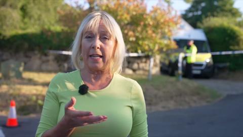 Reporter wearing green top outside Ann Widdecombe's house where a police van is seen and police cordon