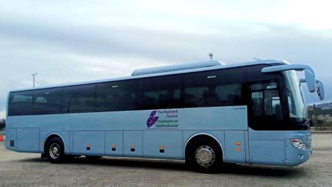 A bus in Highland Council white and purple colours is parked on a large area of hard standing gravel. Some houses and lampposts can be seen in the background. 