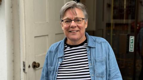 Mary Hervé is smiling as she stands in front of the door of a building. She is wearing a black and white striped top and a denim shirt. She has short grey hair and is wearing glasses.