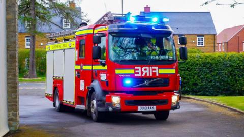 A new-looking fire engine, bright red in colour, faces the camera at a slight angle. Emergency lights glow blue at the top and front of the vehicle. Behind the engine you can see greenery and some residential homes. 