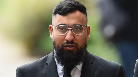 A mid shot of Muhammad Amaad walking towards the camera. He has short dark hair, a long dark beard and is wearing glasses and a black suit and tie.