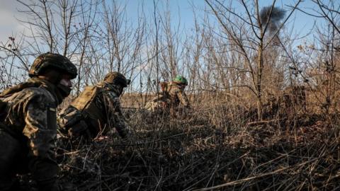 Three Ukrainian soldiers crawling through bushes with an explosion in the distance