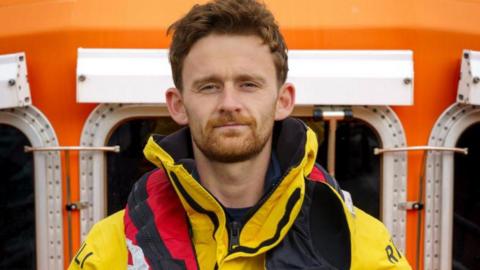 A man with ginger-coloured hair and beard wearing a yellow coat and a red life jacket.