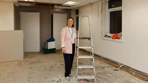 A woman standing in empty room, next to a stepladder. She is wearing dark trousers and a pink and white patterned jacked. The room is unfurnished, and in a state of disrepair, with torn up skirting boards and uncovered and unpainted walls.