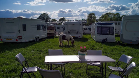 A field with a table and chairs in the foreground and a horse and caravans in the background.
