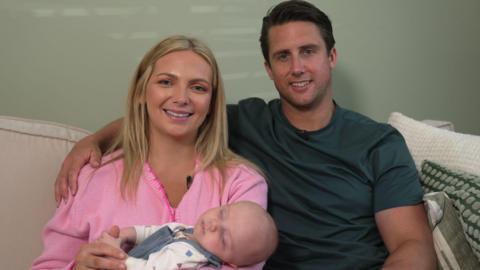 Sleeping baby in the foreground, wearing a white long-sleeved top and blue dungarees, resting across the lap of a seated adult in a pink knitted cardigan. Another seated adult in a dark green short-sleeved top sits alongside with an arm resting behind the first adult. Both adults are seated on a light-coloured sofa with patterned cushions. Small clip-on microphones are attached to the adults’ clothing. A plain light-coloured wall forms the background.