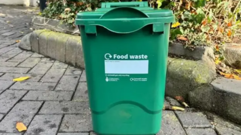 A green food waste caddy on a pavement. It has a recycling logo in white, and the text 'food waste' on the front.