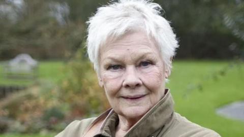 Dame Judi Dench wearing a beige coat and stood outside in front of grass