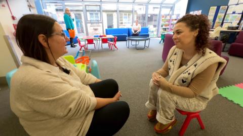 The actor and volunteer sit across from each other on two child-sized chairs in a room laid out with toys and activities for young families.