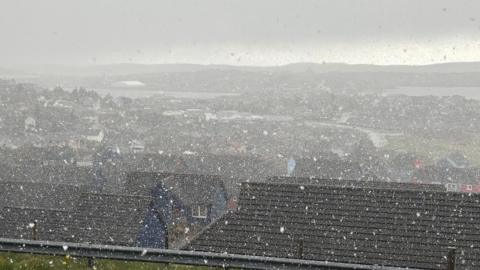 Snow falling across Lerwick in Shetland. Looking out from the top of a hill over lots of houses with black roofs. The sea and hills can be seen in the distance. A dark sky. 