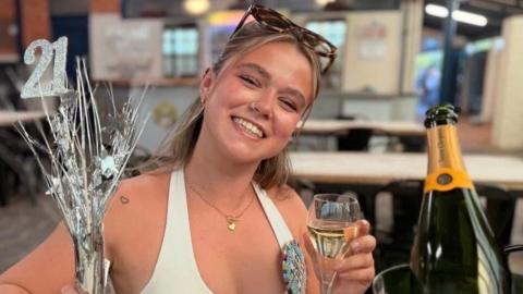 Annie, smiling, holding a glass of champagne in her left hand and a silver 21 birthday decoration in her right hand. She wears a birthday badge, white halterneck top and glasses on top of her head. She is sitting in a restaurant.