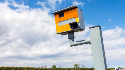 A yellow speed camera box elevated by a pole, pointed at a road in the countryside.