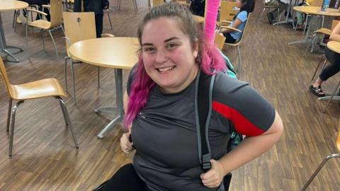 A girl with pink dipped hair holding a back pack whilst she sits in her wheelchair at a table in a cafe.