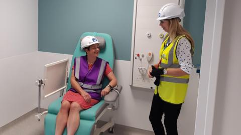 Two women wearing hard hats in the new emergency care centre. One is trying out a patient chair.