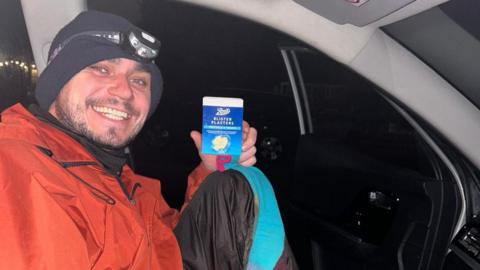 A man in a winter, waterproof jacket smiles sat in the front of a car holding some plasters