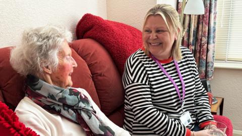 Two people sit on a sofa side by side, a middle aged woman in a black and white striped top and an elderly woman in a white jumper and silk scarf. They are chatting and laughing.