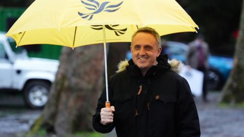 Alex Cole-Hamilton, who has short grey hair, smiles while holding a yellow umbrella