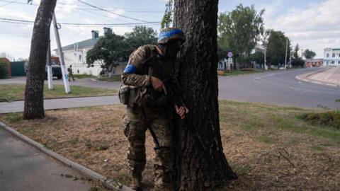A Ukrainian soldier fighting in Russia's Kursk border region. He is carrying an assault rifle and is hiding behind a tree, while wearing a helmet and military fatigues. 
