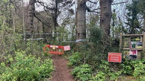 An area of green shrubbery and tall trees with a mud path through the center of it, now fenced off with metal fences and a bright orange fence, with red sign which reads 'footpath ahead closed'.