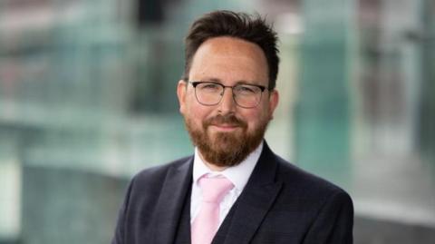 A man with dark hair, glasses and a beard wearing a suit. 