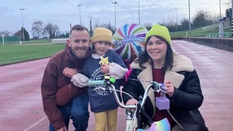 Left to right: Kyle, wearing a brown coat with brown hair and beard kneeling down on an athletic track, with Bobbie wearing a yellow woolly hat, blue jumper and yellow leggings with her scooter and multi-coloured helmet hanging on the handlebars. Her mum Jordan knees down wearing a green woolly hat winter coat and long brown hair. All three are smiling with a congratulations balloon in the background.