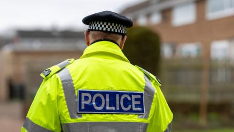 The back of a male police officer wearing a hi-vis jacket which has a police label on the back and he is wearing a black hat