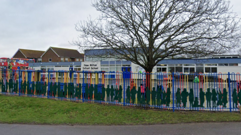 The outside of New Milton Infant School, several buildings behind a blue, red and yellow fence.