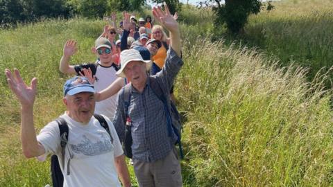 Walkers in a line wave at the camera. They are standing on a footpath running through a field surrounded by grass