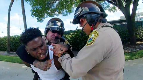 One officer with his arm around the neck of a man who appears to be grimacing, while another officer has his hand on the man's shoulder 