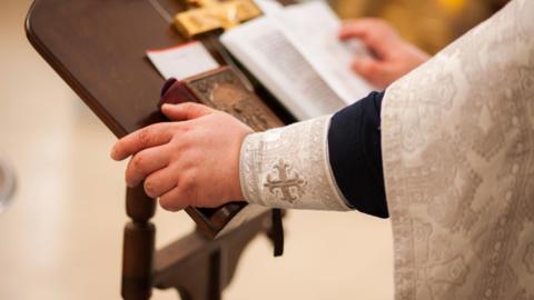 A priest, wearing full vestments, reads from a Bible at a lectern in a church.