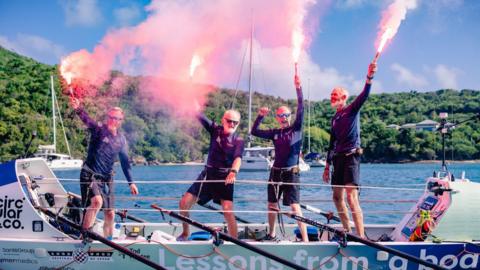 Four men in matching clothes - navy long-sleeve tops and black knee-length shorts. They are in their fifties and sixties. They are stood on a large rowboat which has 'Lessons from a Boat' printed along its side. It is a sunny day, on the coast of Antigua. There are lush trees in the background. All the men are smiling or cheering, with their arms up, holding red flares.