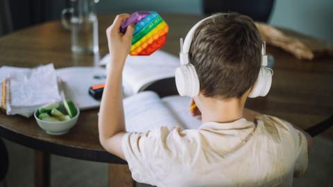 A child sits at a table facing away from the camera. He appears to be doing some school work. He is wearing a pair of white over ear headphones and in his left hand is a multicoloured fidget toy. 