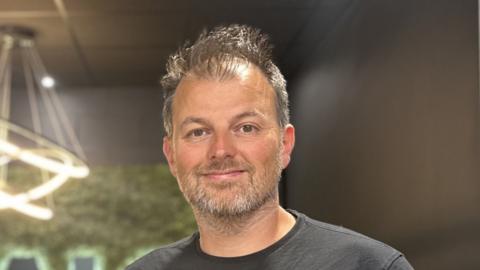 Paul Hamilton smiles at the camera. He has thinning hair that is dark along with a dark beard with some grey hairs in it. He wears a dark T-shirt.