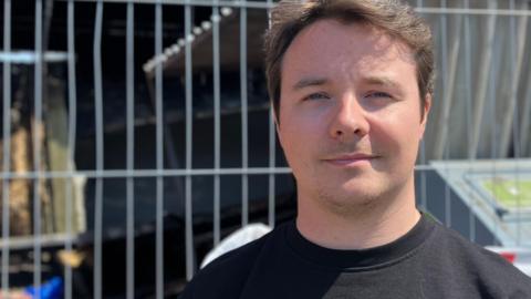 Ryan, a man, with short blonde hair. He is wearing a black T-shirt, smiling for the camera.