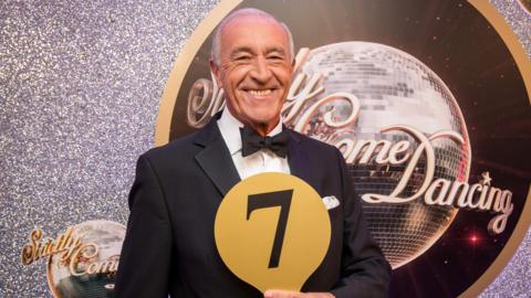 Len Goodman, holding a number seven scorecard, in front of a Strictly Come Dancing logo