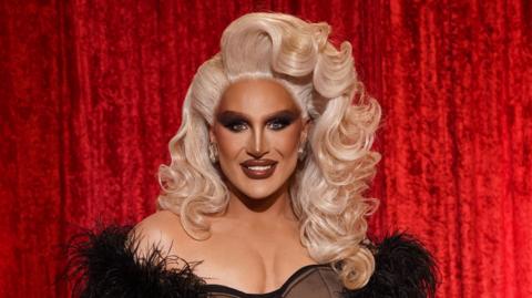 The Vivienne has a large blonde wig on and an open feathery black dress on. The performer is stood in front of a red velvet curtain.