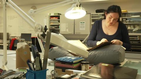 A woman in an office looking through an old ledger with an overhead light over it.
