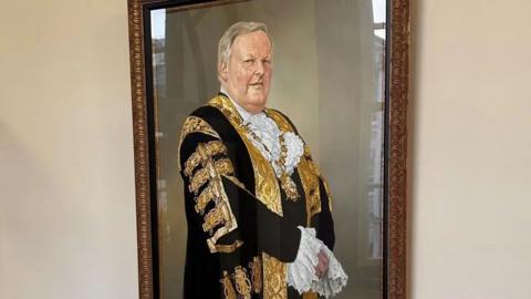 The portrait of former Belfast lord mayor Lord Browne on display again at City Hall. Lord Browne is wearing black and gold official robes and his gold chain of office. His hands are clasped in front of him.