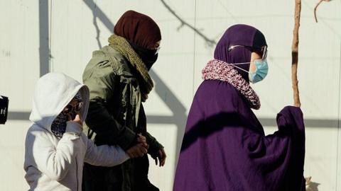 A side profile of two women in Islamic dress and scarves and a child in a grey hoodie and sunglasses.