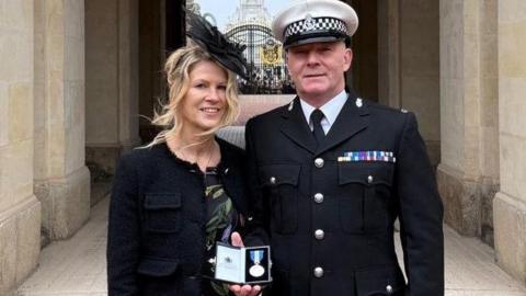 A man stands with his wife in front of an archway at Buckingham Palace, holding a medal, held in a box, in his right hand. He is dressed in formal police uniform, while she is wearing a flowery dress and a black hat, with a black jacket 