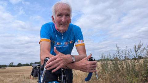 An older man with white hair is on a bicycle in a field of crops. He is wearing a blue cycling t-shirt that reads "hope house children's hospices"