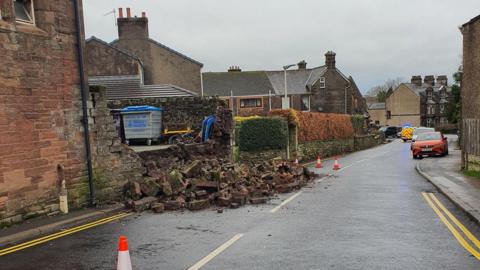 Bricks from the collapsed wall are in the middle of the street, surrounded by orange high-viz cones. The collapsed wall appears to be from the school's yard. A police van is blocking the road