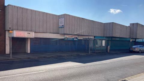 Empty shop units along Osborne Street. They have flat concrete roofs and windows with blue metal shutters in front of them